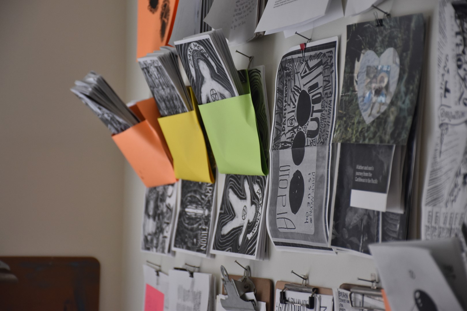 A wall shot in a depth of field blur by a wall of zines and paper held hostage in clipboards